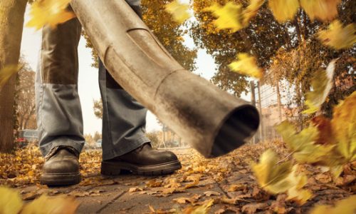 A person is using a leaf blower to clean up a sidewalk in the city. Colorful leaves are swirling around. Low perspective showing the ground and shoes.