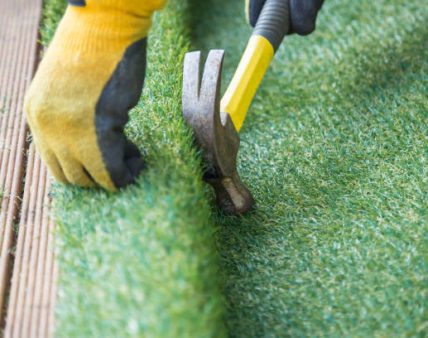 Artificial grass, turf installation alongside decking. A hammer in being used to nail the turf into place. A yellow work glove can be seen.